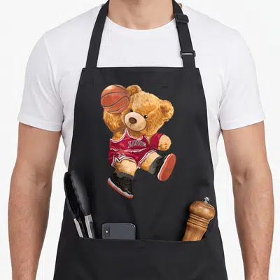 Playing teddy bear apron - Brown teddy bear in red jersey holding orange basketball with LOVE text — shown on a black cotton canvas apron against a white background