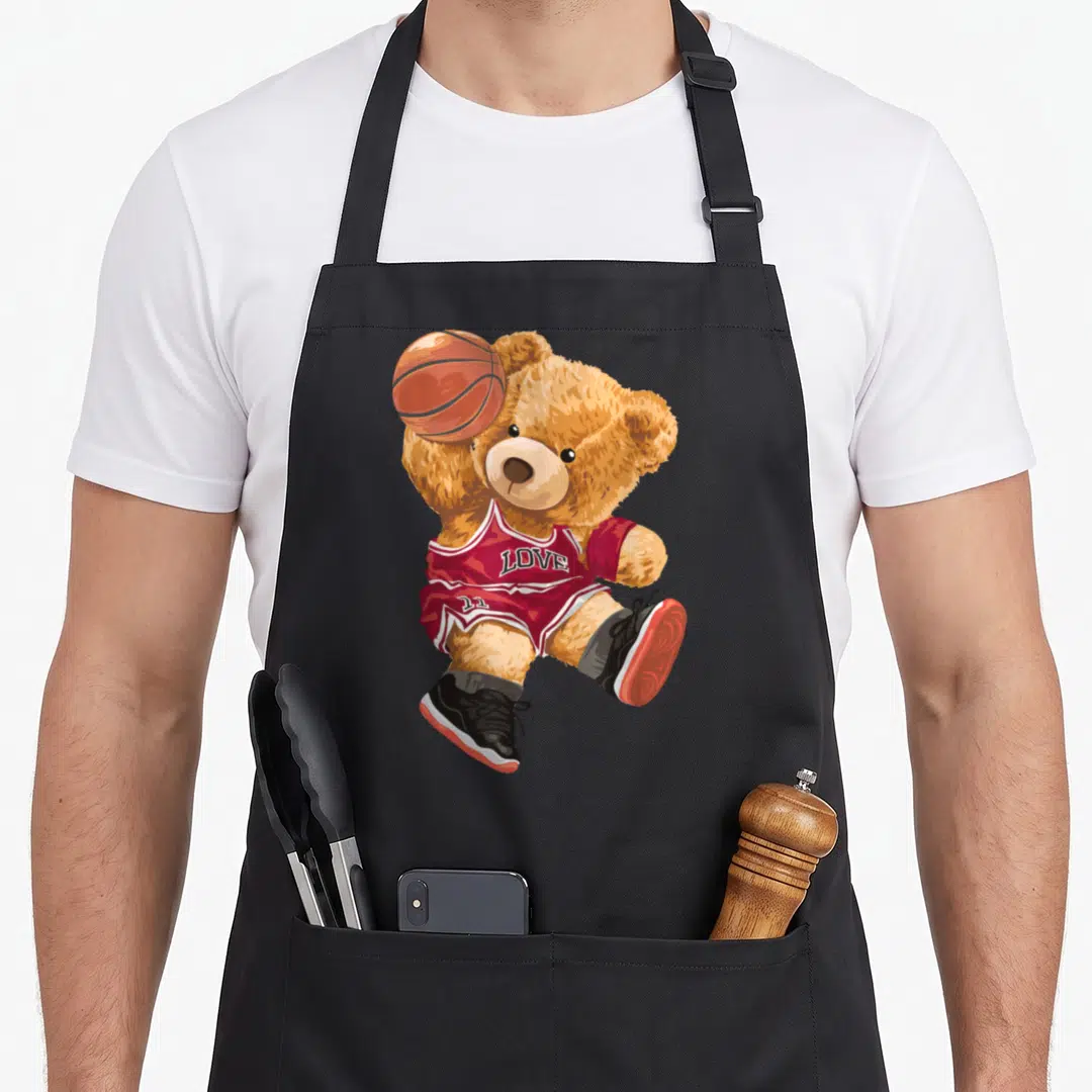 Playing teddy bear apron - Brown teddy bear in red jersey holding orange basketball with LOVE text — shown on a black cotton canvas apron against a white background