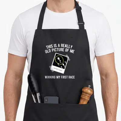 Race gag aprons - Sperm cell photo with winner's circle text and yellow highlight on taped picture — shown on a black cotton canvas apron against a white background