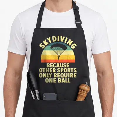 Skydiving sky apron - Retro rainbow skydiver silhouette with text about requiring one ball for parachuting — shown on a black cotton canvas apron against a white background