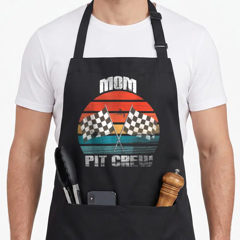 Chekered crew aprons - MOM PIT CREW text with two crossed checkered flags over a retro striped sun — shown on a black cotton canvas apron against a white background