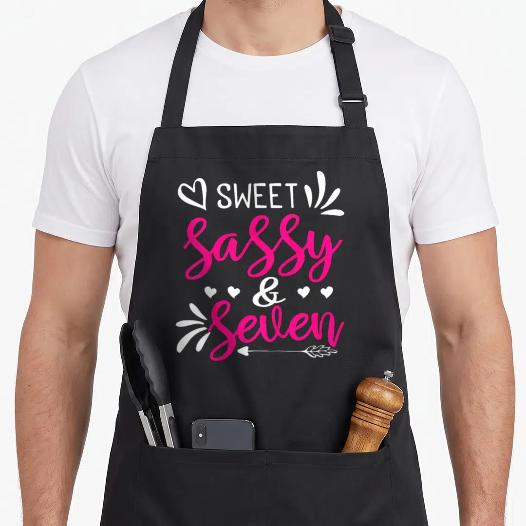Bday sassy apron - Stacked SWEET Sassy & Seven text with hearts and arrow in white and pink — shown on a black cotton canvas apron against a white background
