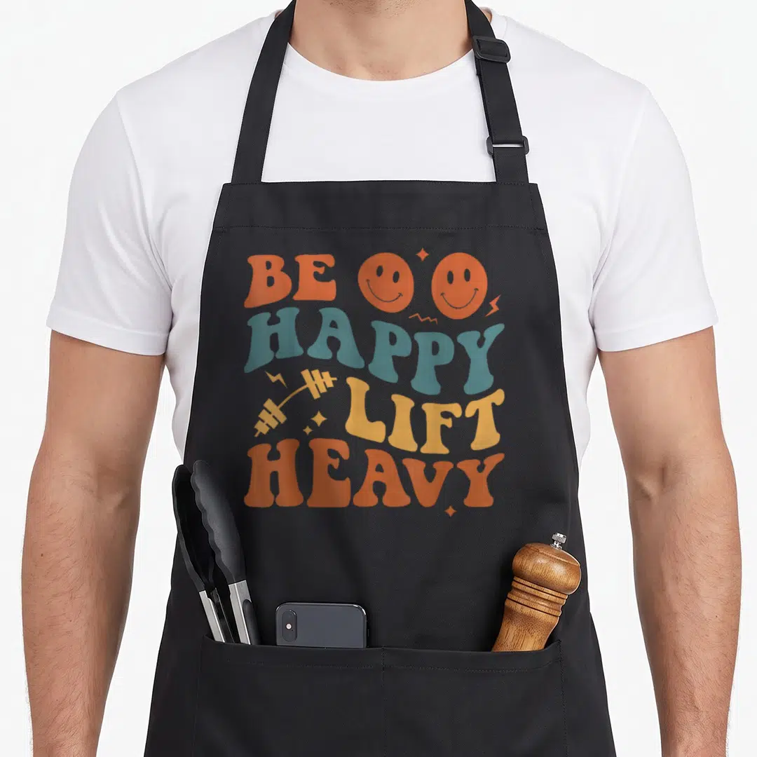Workout heavy apron - Retro text BE HAPPY LIFT HEAVY with orange smiley faces and yellow barbell on four lines — shown on a black cotton canvas apron against a white background