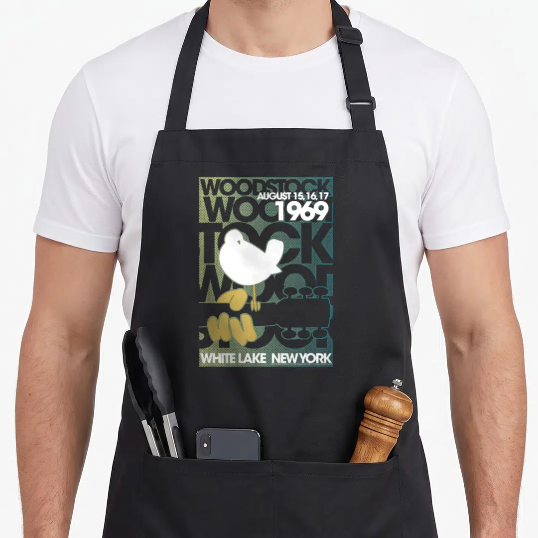 Lake woodstock era apron - White bird perched on yellow-green guitar with WOODSTOCK text and 1969 dates on blue-grey — shown on a black cotton canvas apron against a white background