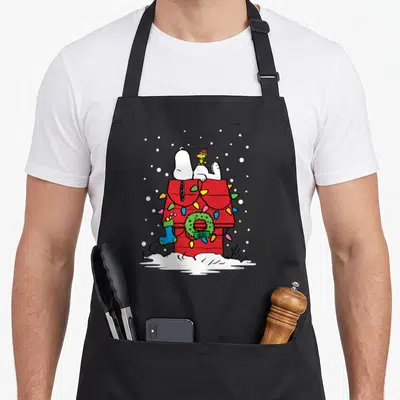 Woodstock christmas stocking apron - Snoopy and Woodstock on red doghouse with green wreath, blue stocking — shown on a black cotton canvas apron against a white background