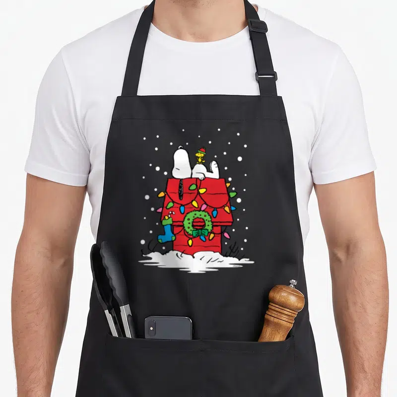 Woodstock christmas stocking apron - Snoopy and Woodstock on red doghouse with green wreath, blue stocking — shown on a black cotton canvas apron against a white background