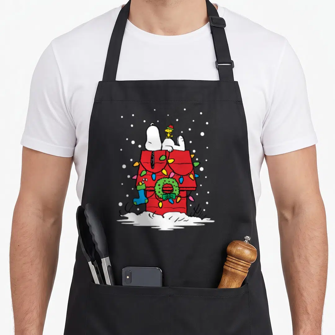 Woodstock christmas stocking apron - Snoopy and Woodstock on red doghouse with green wreath, blue stocking — shown on a black cotton canvas apron against a white background