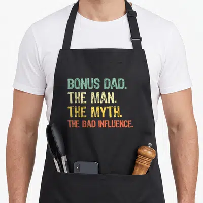 Myth bonus dad apron - Stacked text reading BONUS DAD THE MAN THE MYTH THE BAD INFLUENCE in green beige yellow — shown on a black cotton canvas apron against a white background