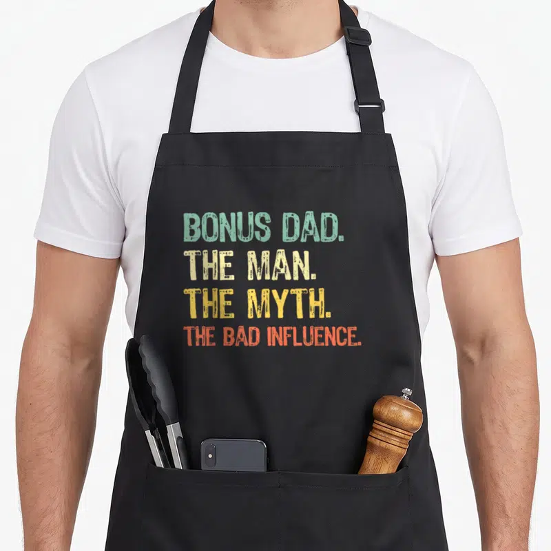 Myth bonus dad apron - Stacked text reading BONUS DAD THE MAN THE MYTH THE BAD INFLUENCE in green beige yellow — shown on a black cotton canvas apron against a white background