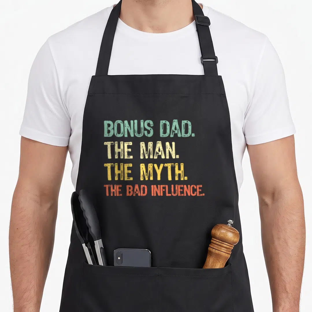 Myth bonus dad apron - Stacked text reading BONUS DAD THE MAN THE MYTH THE BAD INFLUENCE in green beige yellow — shown on a black cotton canvas apron against a white background