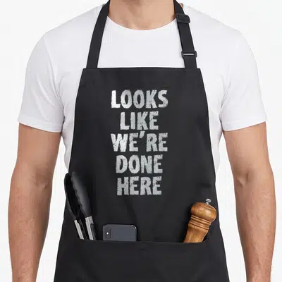 Sweat drenched apron - White stacked text reading Looks Like We're Done Here in a distressed font style — shown on a black cotton canvas apron against a white background
