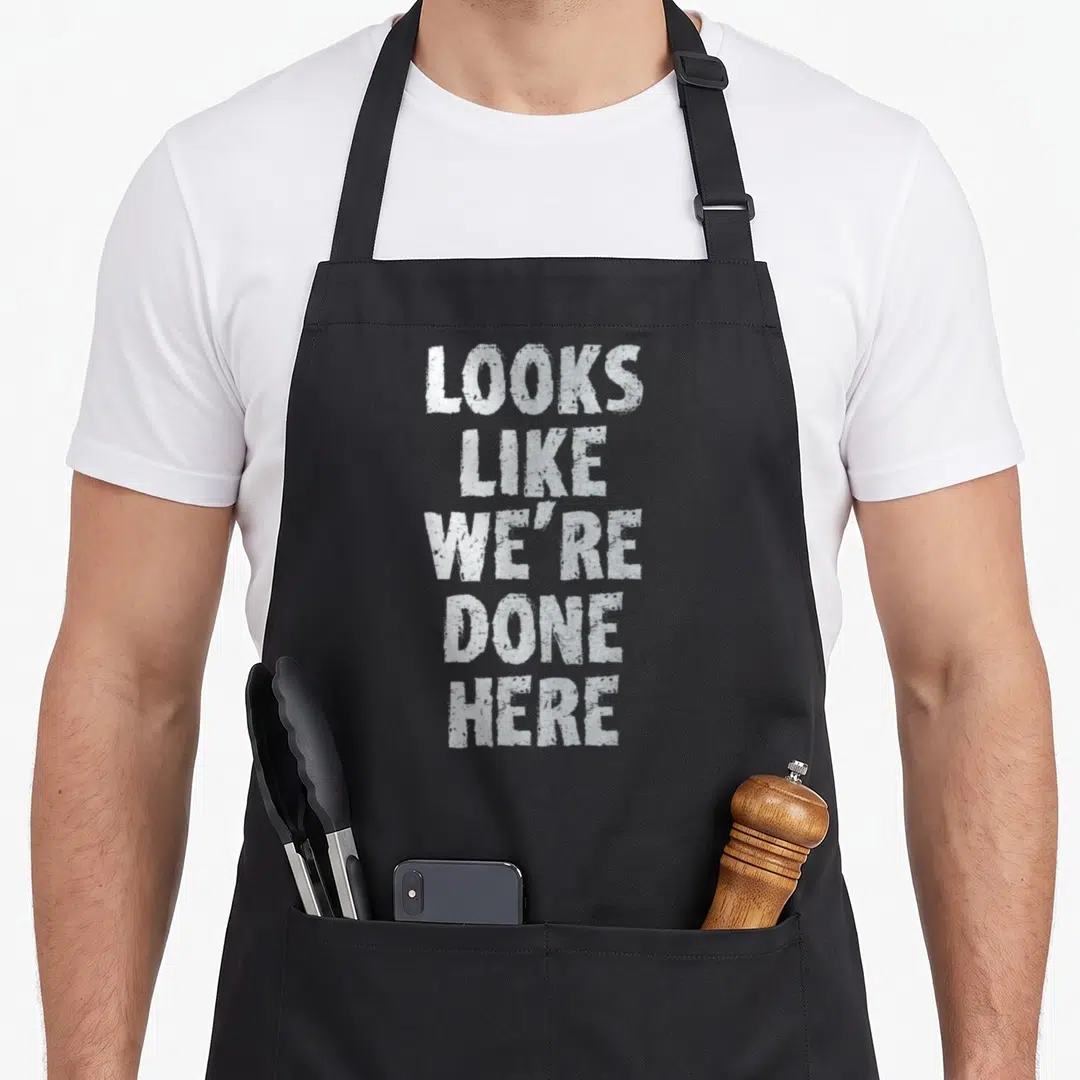 Sweat drenched apron - White stacked text reading Looks Like We're Done Here in a distressed font style — shown on a black cotton canvas apron against a white background