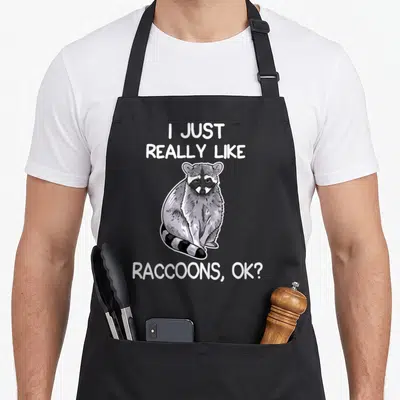 Vintage raccoon apron - Cartoon raccoon centered between text I Just Really Like Raccoons OK in white, grey, black — shown on a black cotton canvas apron against a white background