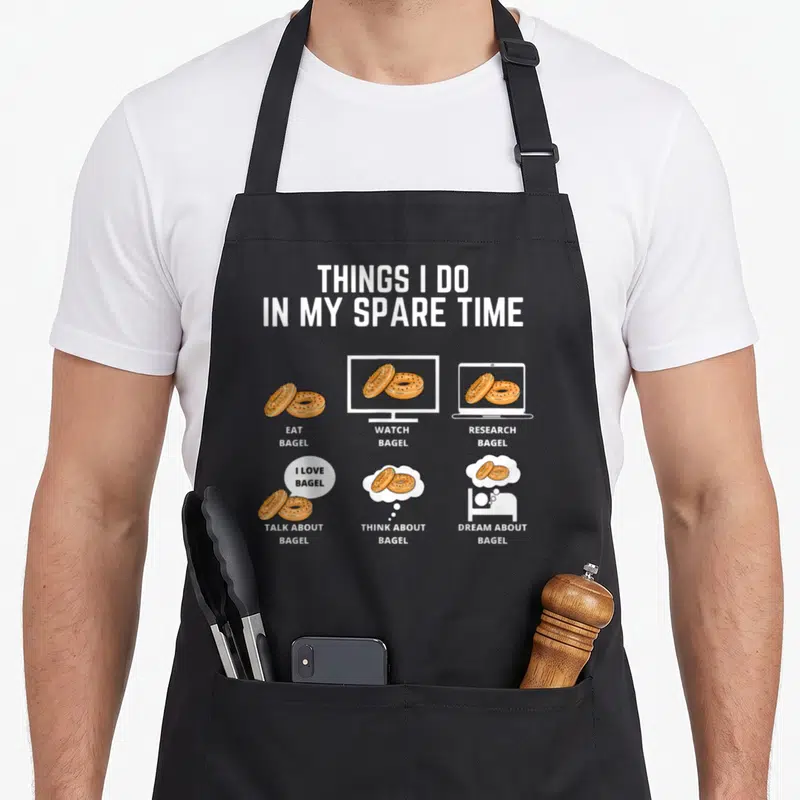 Bagel spare time apron - White text THINGS I DO IN MY SPARE TIME above six cartoon bagel icons in orange and brown — shown on a black cotton canvas apron against a white background