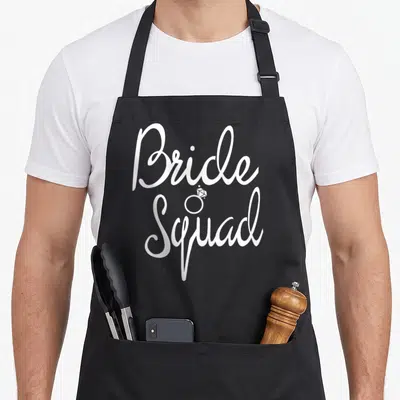 Bride bridesmaid aprons - White Bride Squad script with diamond ring icon centered between words — shown on a black cotton canvas apron against a white background