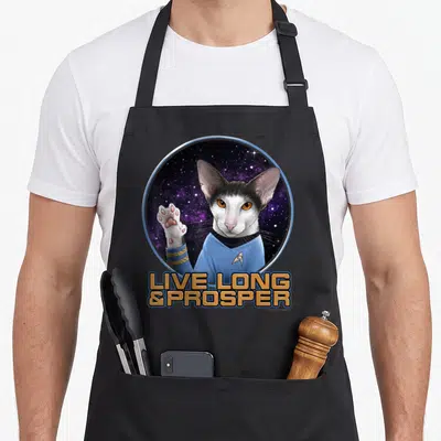 Original cat apron - Realistic cat in blue uniform doing Vulcan salute with LIVE LONG & PROSPER text — shown on a black cotton canvas apron against a white background