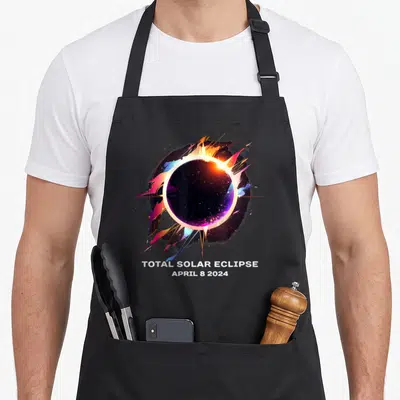 Souvenir april apron - Total Solar Eclipse April 8 2024 text below colorful circular eclipse graphic with orange pink — shown on a black cotton canvas apron against a white background