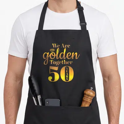 Together 50th wedding aprons - Golden stacked text We Are golden Together 50 YEARS with diamond icon — shown on a black cotton canvas apron against a white background