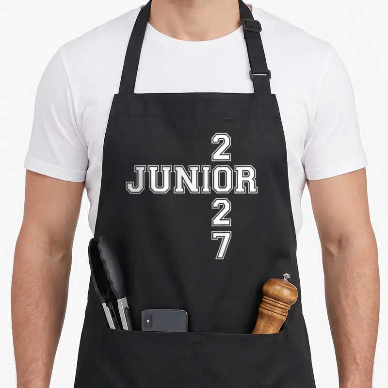 Student junior apron - White 2027 JUNIOR typography in cross layout on black fabric — shown on a black cotton canvas apron against a white background