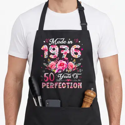 50th vintage floral apron - Stacked Made in 1976 text with floral filled numbers and pink rose bouquet — shown on a black cotton canvas apron against a white background