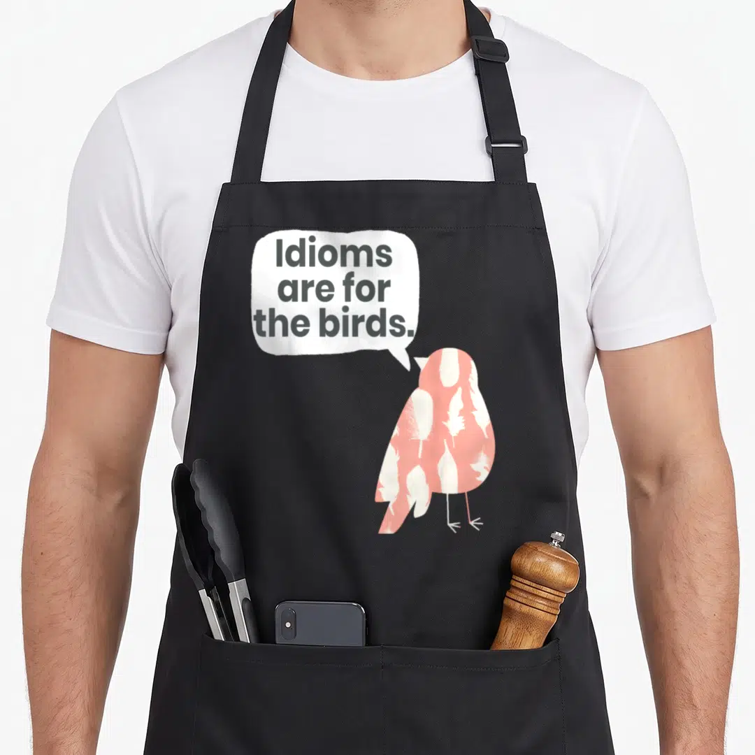 Idiom teacher apron - Pink bird with white feathers and speech bubble reading Idioms are for the birds on grey — shown on a black cotton canvas apron against a white background