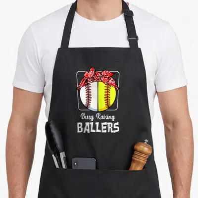 Softball busy apron - Split baseball with red bandana and Busy Raising Ballers text — shown on a black cotton canvas apron against a white background