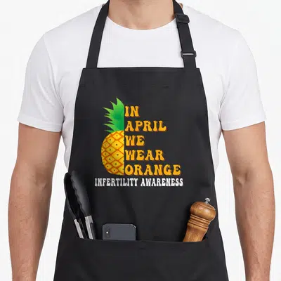 Week april apron - Half pineapple with stacked orange text reading IN APRIL WE WEAR ORANGE and white text below — shown on a black cotton canvas apron against a white background
