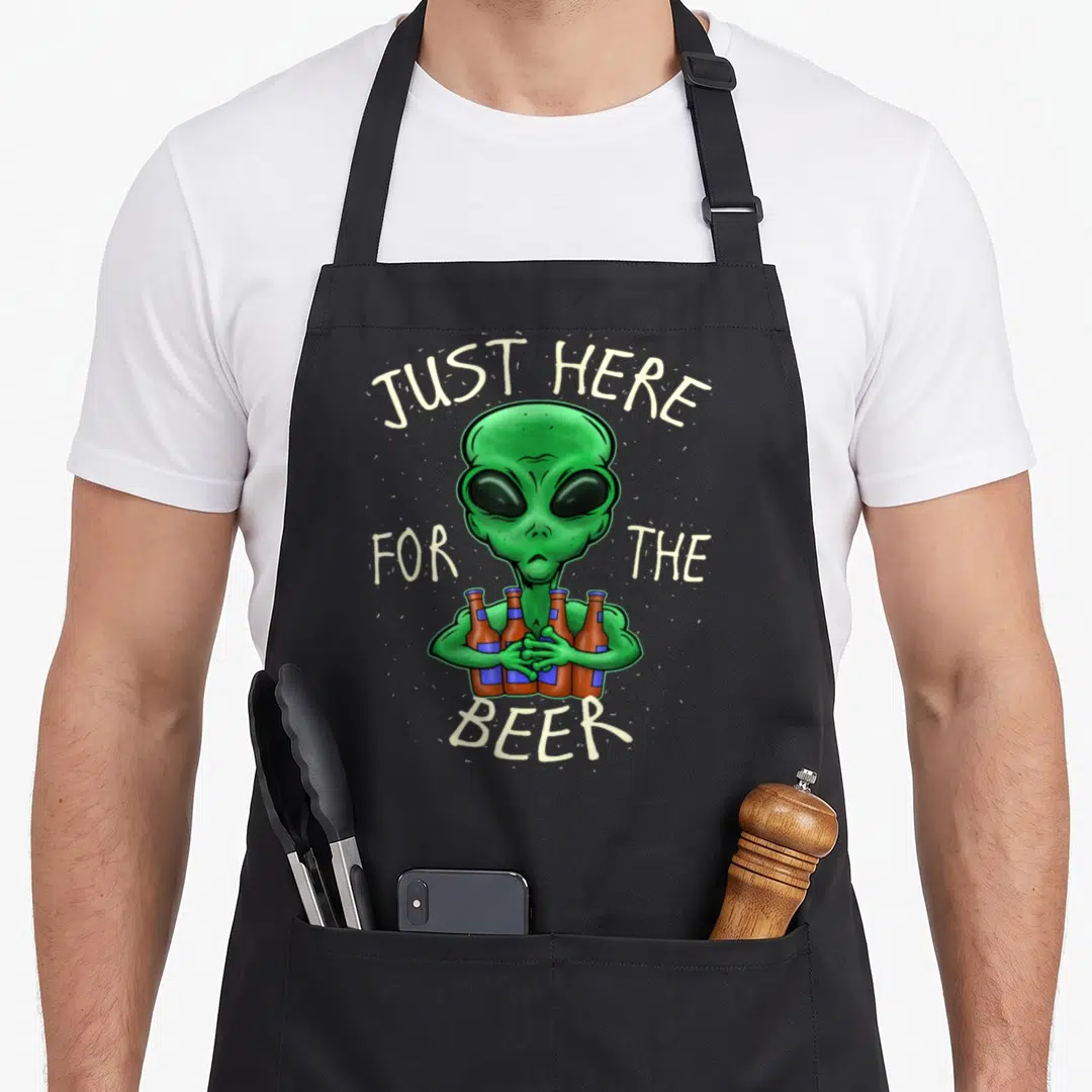Alien beer apron - Green cartoon alien holding four brown beer bottles with arched text JUST HERE FOR THE BEER — shown on a black cotton canvas apron against a white background