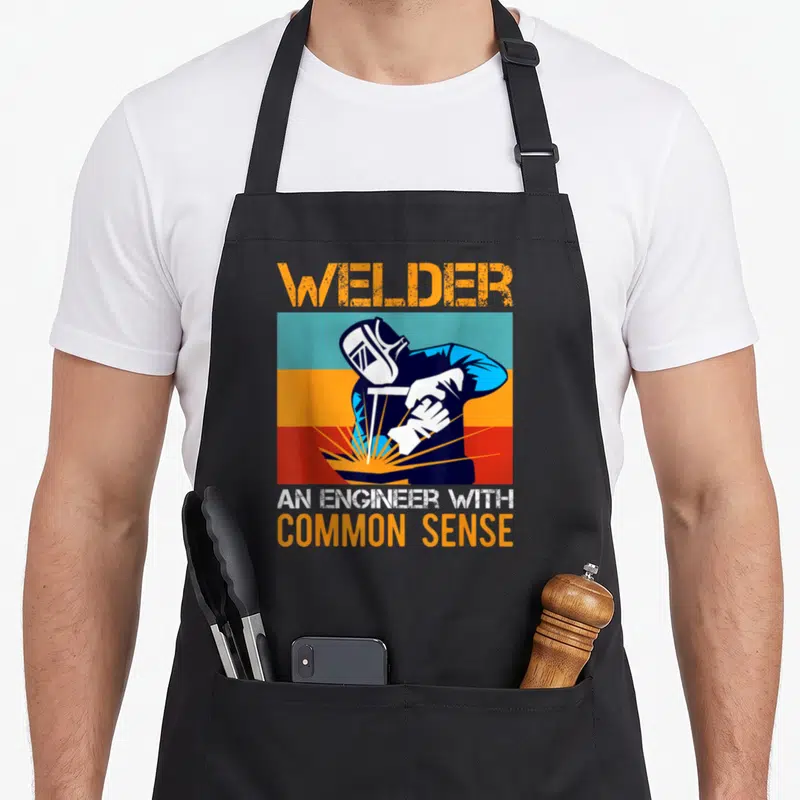 Cute welding welder dad apron - Retro graphic of welder in mask with horizontal orange blue teal red stripes and text — shown on a black cotton canvas apron against a white background