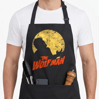 Wolf ogre apron - Black werewolf silhouette against yellow moon with red distressed THE WOLFMAN text — shown on a black cotton canvas apron against a white background