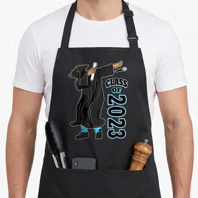 Senior matter grad apron - Cartoon graduate in black robe dabbing with diploma and vertical Class of 2023 text — shown on a black cotton canvas apron against a white background