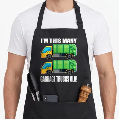 Garbage 2nd birthday truck apron - Two stacked yellow and green cartoon garbage trucks with white text reading I'M THIS — shown on a black cotton canvas apron against a white background