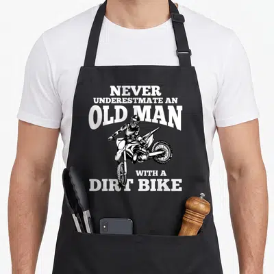 Underestimate bike apron - White text NEVER UNDERESTIMATE AN OLD MAN WITH A DIRT BIKE around black line-art dirt bike — shown on a black cotton canvas apron against a white background