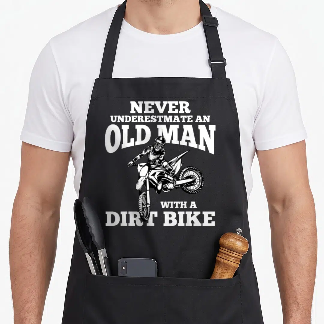Underestimate bike apron - White text NEVER UNDERESTIMATE AN OLD MAN WITH A DIRT BIKE around black line-art dirt bike — shown on a black cotton canvas apron against a white background
