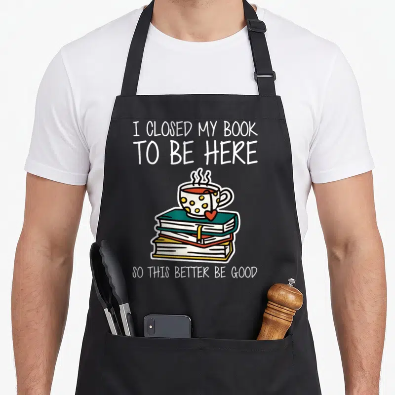 Closed better apron - I closed my book to be here so this better be good text with coffee cup on books — shown on a black cotton canvas apron against a white background
