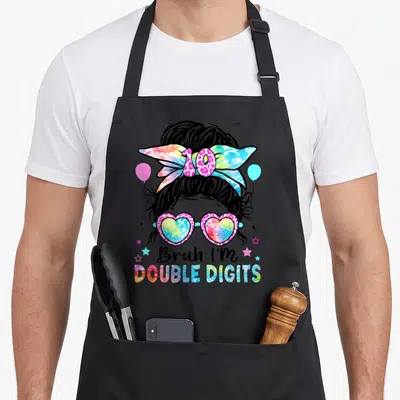 Bruh double digit 10th birthday apron - Cartoon messy bun girl with heart glasses, tie-dye bow, and stacked text — shown on a black cotton canvas apron against a white background