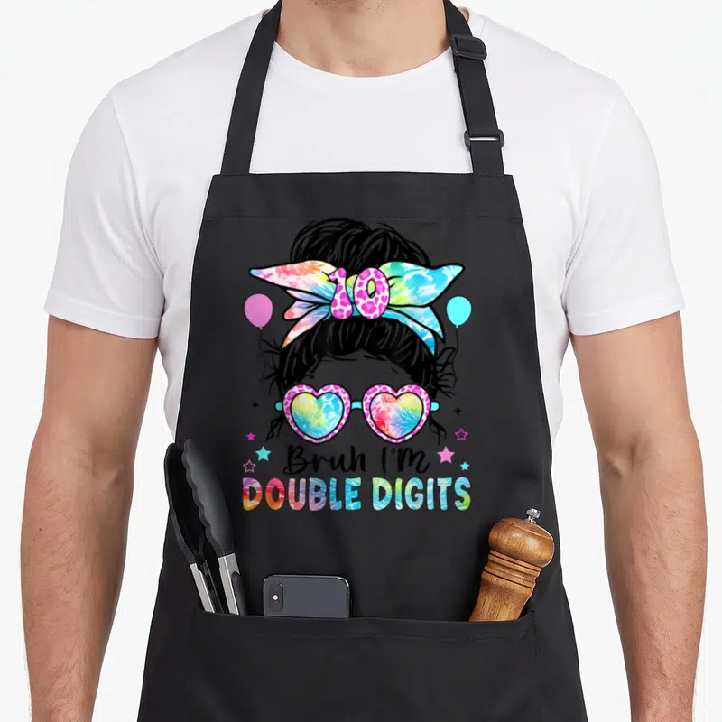 Bruh double digit 10th birthday apron - Cartoon messy bun girl with heart glasses, tie-dye bow, and stacked text — shown on a black cotton canvas apron against a white background