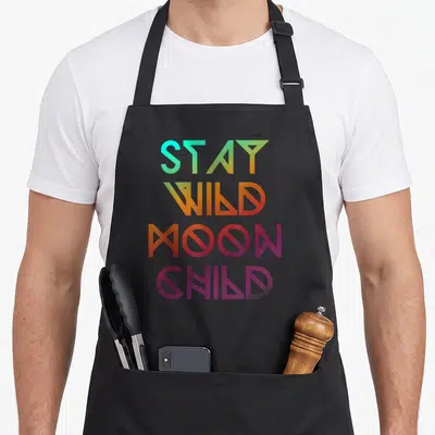 Wicca moon child apron - STAY WILD MOON CHILD text in cyan orange purple pink geometric font — shown on a black cotton canvas apron against a white background