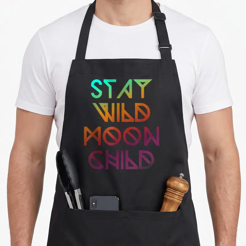 Wicca moon child apron - STAY WILD MOON CHILD text in cyan orange purple pink geometric font — shown on a black cotton canvas apron against a white background