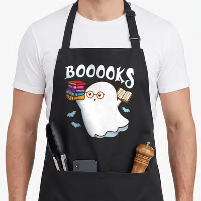Book halloween apron - Ghost with brown glasses holding books under arched BOOOOKS text with two blue bats — shown on a black cotton canvas apron against a white background