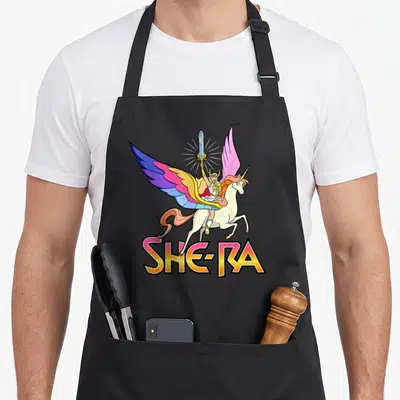 Power princess apron - She-Ra riding a winged unicorn holding a sword above gradient SHE-RA text — shown on a black cotton canvas apron against a white background