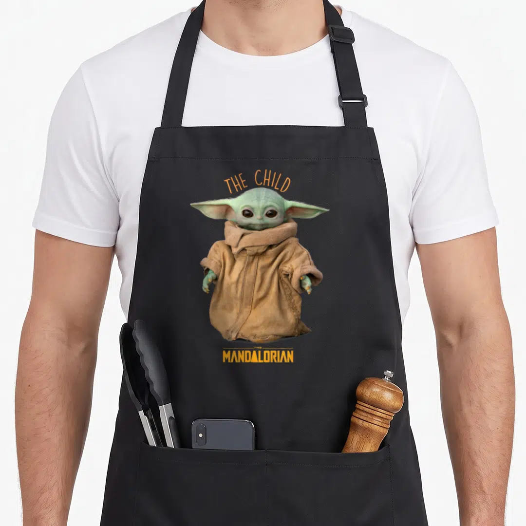 Pose star war apron - Photo of Grogu in tan robe with orange THE CHILD and THE MANDALORIAN text — shown on a black cotton canvas apron against a white background