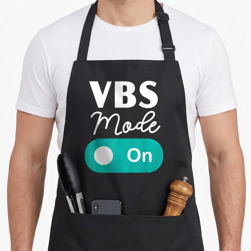 Bible teacher apron - Stacked VBS Mode On text above teal toggle switch graphic — shown on a black cotton canvas apron against a white background
