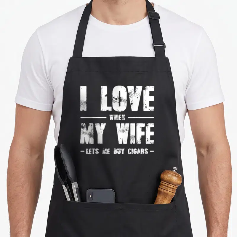Cigar wife apron - White distressed text I LOVE WHEN MY WIFE LETS ME BUY CIGARS stacked layout — shown on a black cotton canvas apron against a white background