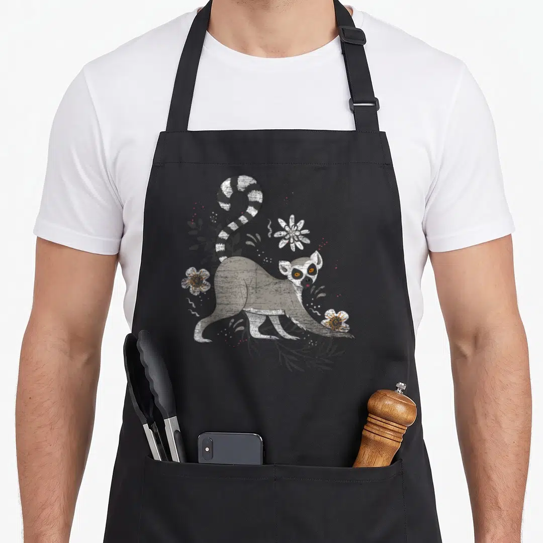 Madagascar animal apron - Cartoon ring-tailed lemur with grey leaves and white flowers with orange centers — shown on a black cotton canvas apron against a white background