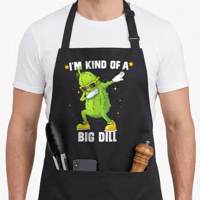 Kind halloween apron - Green pickle character dabbing with sunglasses between white text lines and yellow stars — shown on a black cotton canvas apron against a white background