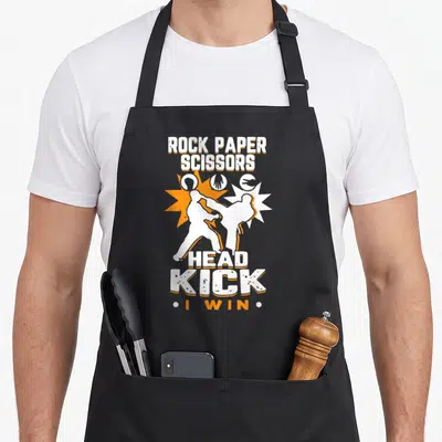 Youth taekwondo apron - Rock Paper Scissors Head Kick I Win text with two fighting silhouettes and hand symbols — shown on a black cotton canvas apron against a white background