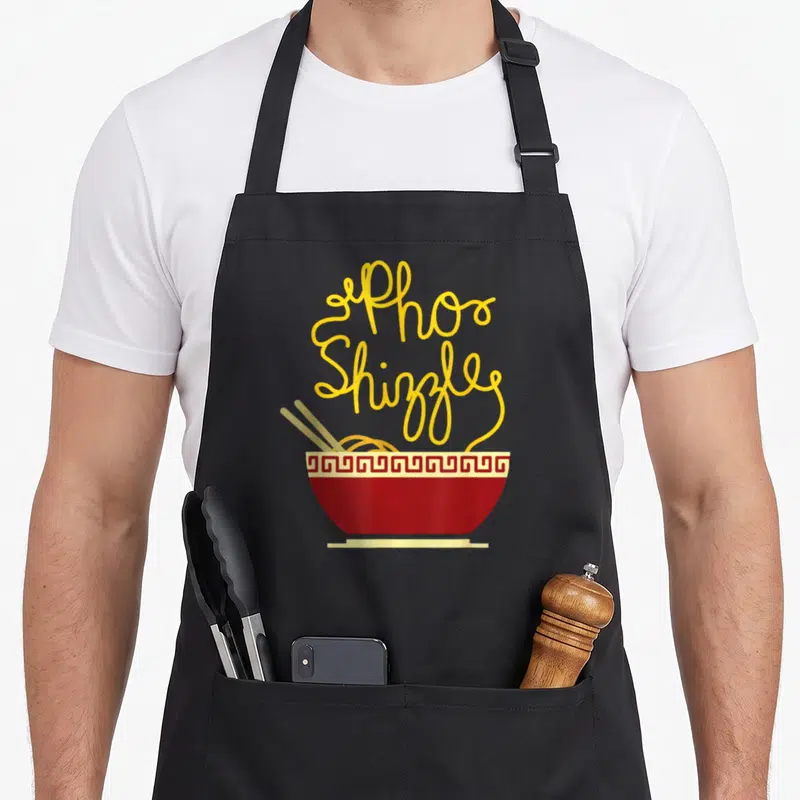 Pho asian apron - Yellow Pho Shizzle noodles rising from red bowl with chopsticks — shown on a black cotton canvas apron against a white background
