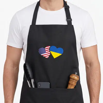 Ukraine ukrainian american flag apron - Two overlapping hearts, one US flag pattern, one blue and yellow Ukrainian flag — shown on a black cotton canvas apron against a white background