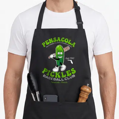 Pensacola pickle apron - Green cartoon pickle holding a brown bat with arching green and white text reading Pensacola — shown on a black cotton canvas apron against a white background